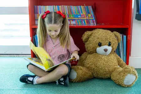 Lower School Girl  in the library reading quietly to a large teddy sitting next to her.